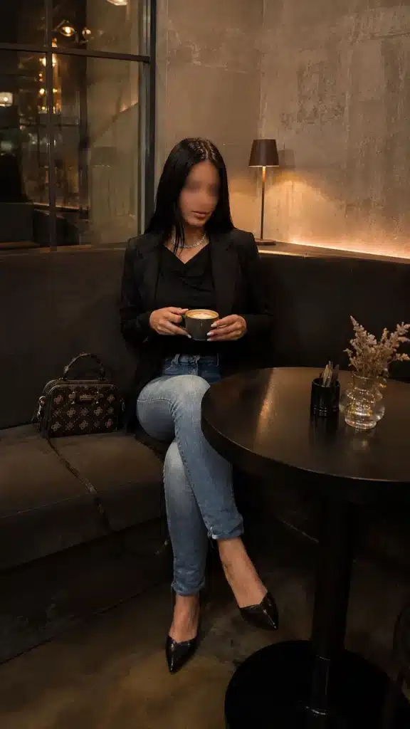 Eine Frau mit langen dunklen Haaren sitzt auf einem dunklen Sofa in einem modernen Café und hält eine Tasse in der Hand. Sie trägt einen schwarzen Blazer, blaue Jeans und schwarze Pumps. Neben ihr steht eine Designer-Handtasche, und auf einem runden Tisch stehen eine Vase und Besteck.