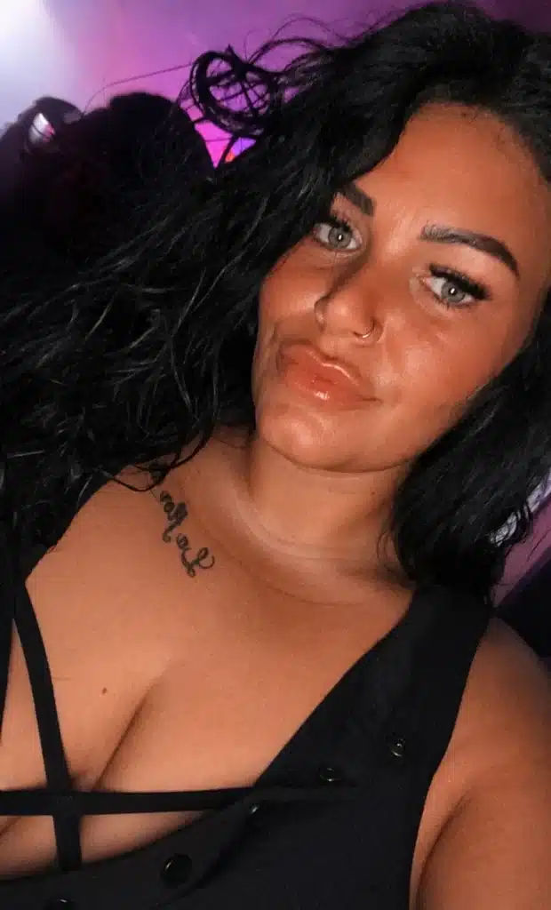 A woman with long dark hair and blue eyes is taking a selfie. She is wearing a black top with crisscrossed straps, has a nose ring and a visible tattoo on her chest, and is standing in a dimly lit setting with purple and pink light in the background.