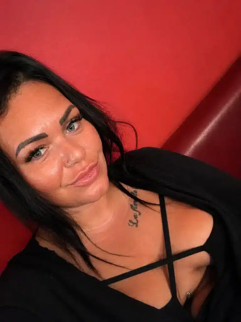 A woman with long dark hair and blue eyes, wearing a black top with crisscrossed straps, is sitting in front of a red background. She has a nose ring and a tattoo on her upper chest.