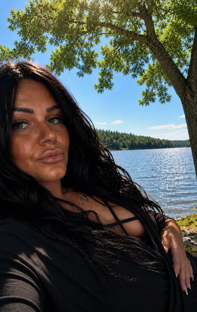 A woman with long dark hair is taking a selfie outside near a lake, while sunlight filters through the green foliage of the trees and a bright blue sky is visible in the background.