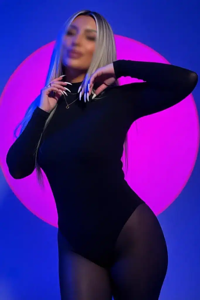 Barbie mit langen blonden Haaren trägt einen schwarzen Bodysuit und posiert mit den Händen im Nacken.