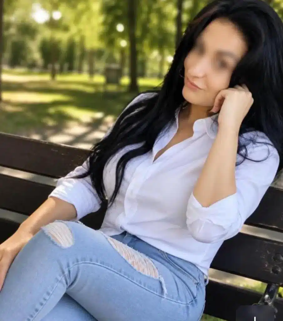 A person with long dark hair, wearing a white shirt and ripped light blue jeans, is sitting on a park bench with one hand resting against their cheek. In the background, you can see trees and a sunlit park.