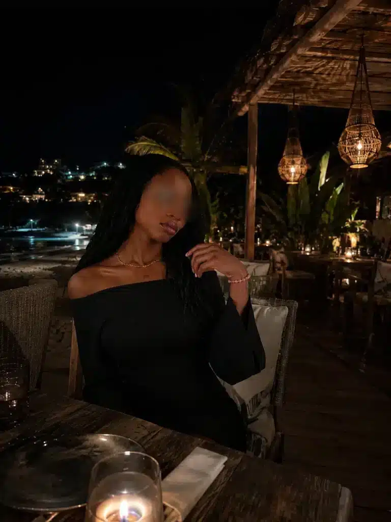A woman in a black off-the-shoulder dress is sitting at night in an outdoor restaurant lit by candles, surrounded by tropical plants and hanging lanterns, with the city lights in the background. Her face is out of focus.