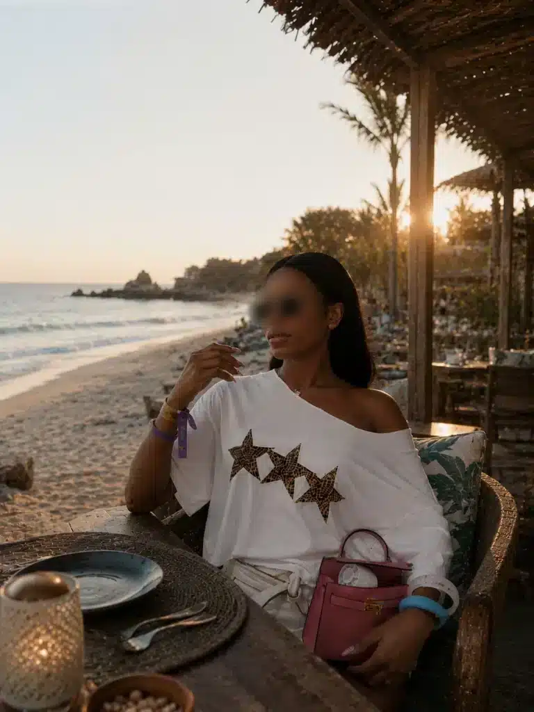 A woman with blurred facial features is sitting in a beach restaurant at sunset. She is wearing a white shirt with a star pattern, holding a glass in her hand, and has a pink bag on her lap. The ocean and palm trees can be seen in the background.