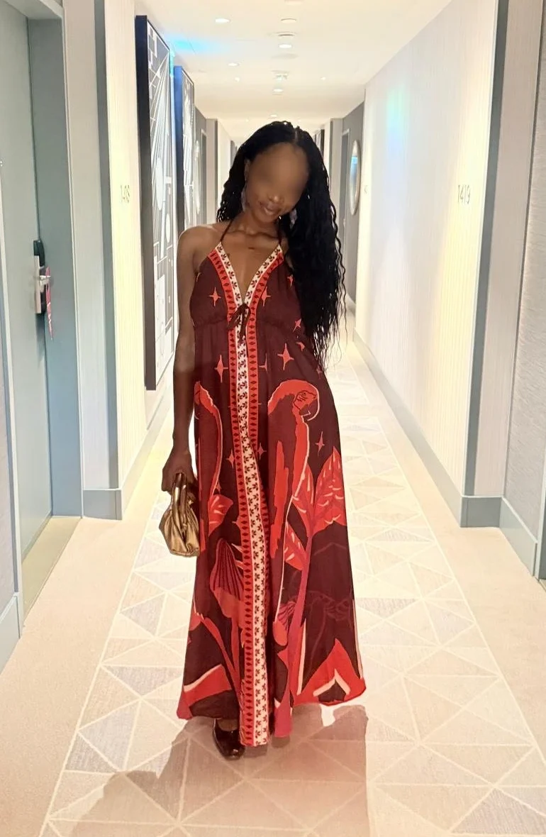 Coco in einem fließenden, rot gemusterten Kleid steht in einem gut beleuchteten Flur mit hellen Wänden.