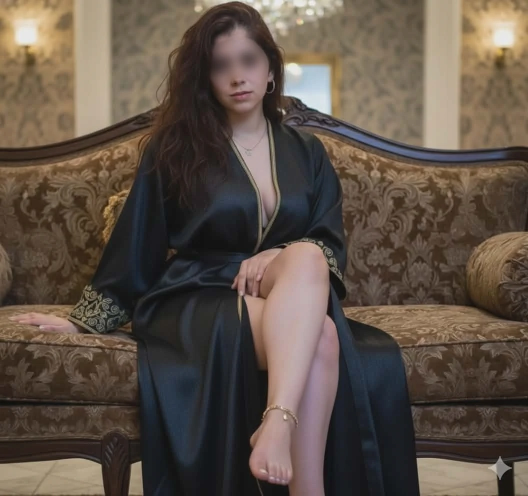 Dalia mit langen dunklen Haaren sitzt auf einem gemusterten Sofa und trägt ein seidiges schwarzes Gewand mit Goldstickerei.