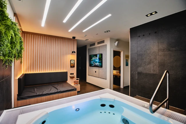 Moderner Spa-Raum mit einem Whirlpool im Vordergrund, einer schwarz gepolsterten Sitzecke mit Holzlatten und warmer Beleuchtung auf der linken Seite, perfekt für Wellness und Entspannung, sowie einem an der Wand montierten Fernseher und einer Tür zum Badezimmer im Hintergrund.