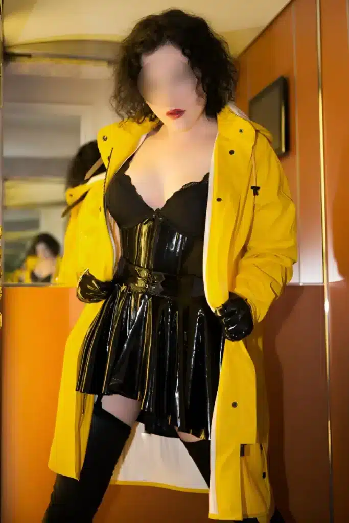 Eine Person mit verschwommenen Gesichtszügen trägt ein glänzendes schwarzes Outfit, schwarze Handschuhe, oberschenkelhohe Stiefel und einen leuchtend gelben Regenmantel und steht in einem Innenraum vor einer verspiegelten Wand.