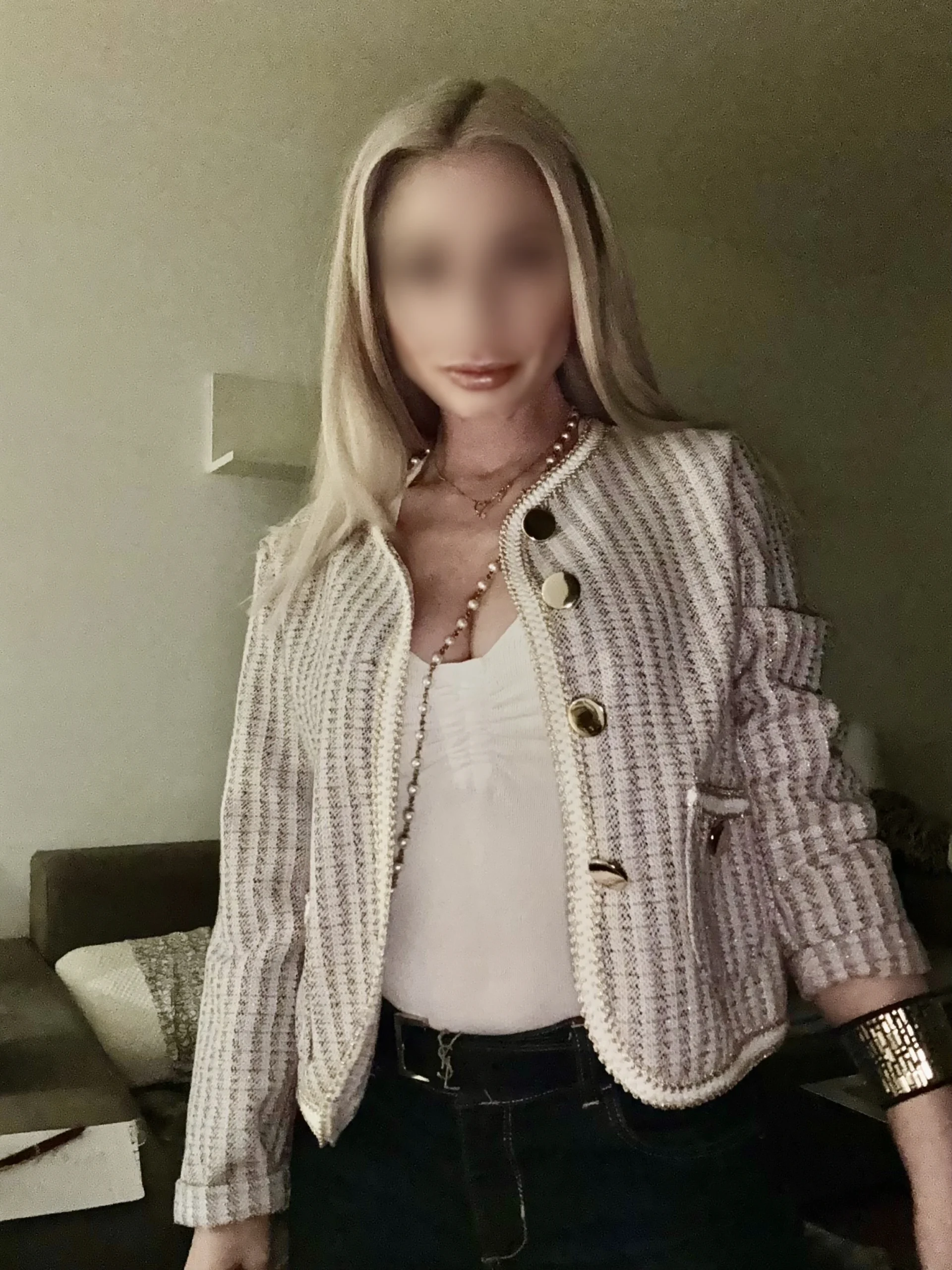 Eine Frau mit langen blonden Haaren, die eine strukturierte helle Jacke mit goldenen Knöpfen über einem weißen Oberteil und einer dunklen Hose trägt, steht in einem Innenraum. Ihr Gesicht ist unscharf, und im Hintergrund sind ein Sofa und ein Tisch zu sehen.