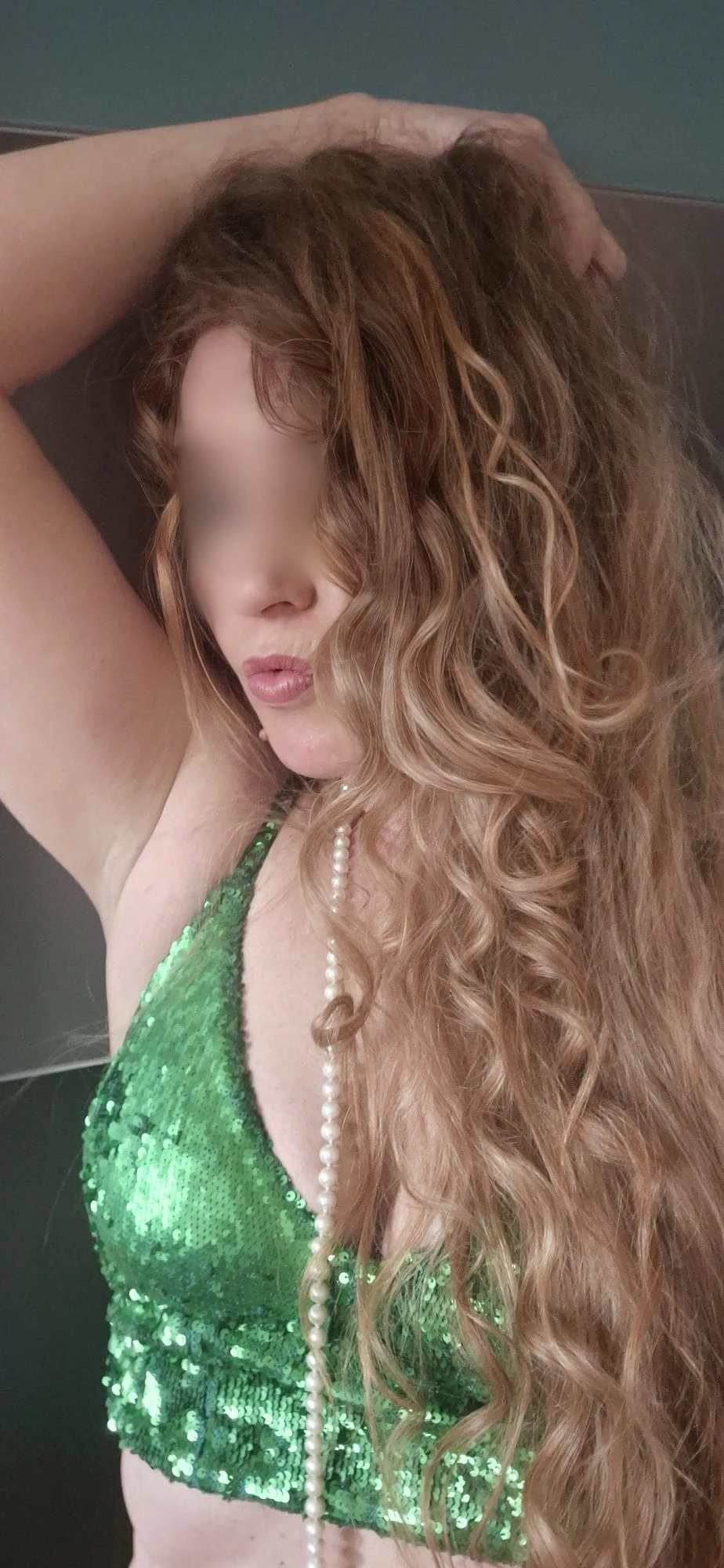 Escort Wien Heidi mit langen, gewellten blonden Haaren, die ein glitzerndes grünes Oberteil trägt.