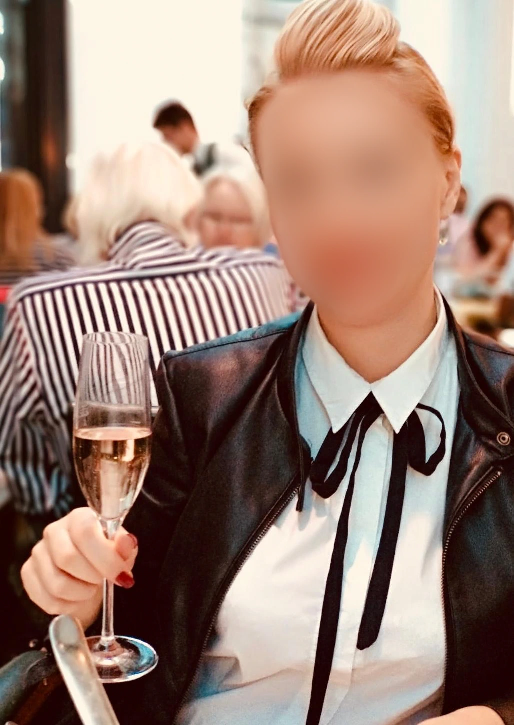 Irina, mit kurzen blonden Haaren, trägt eine schwarze Jacke und ein weißes Hemd mit einer schwarzen Krawatte und hält ein Glas Sekt.