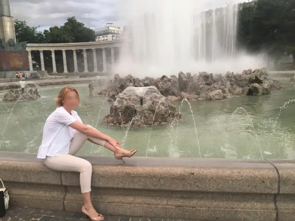 Irina, leicht bekleidet, sitzt am Rande eines steinernen Brunnens.