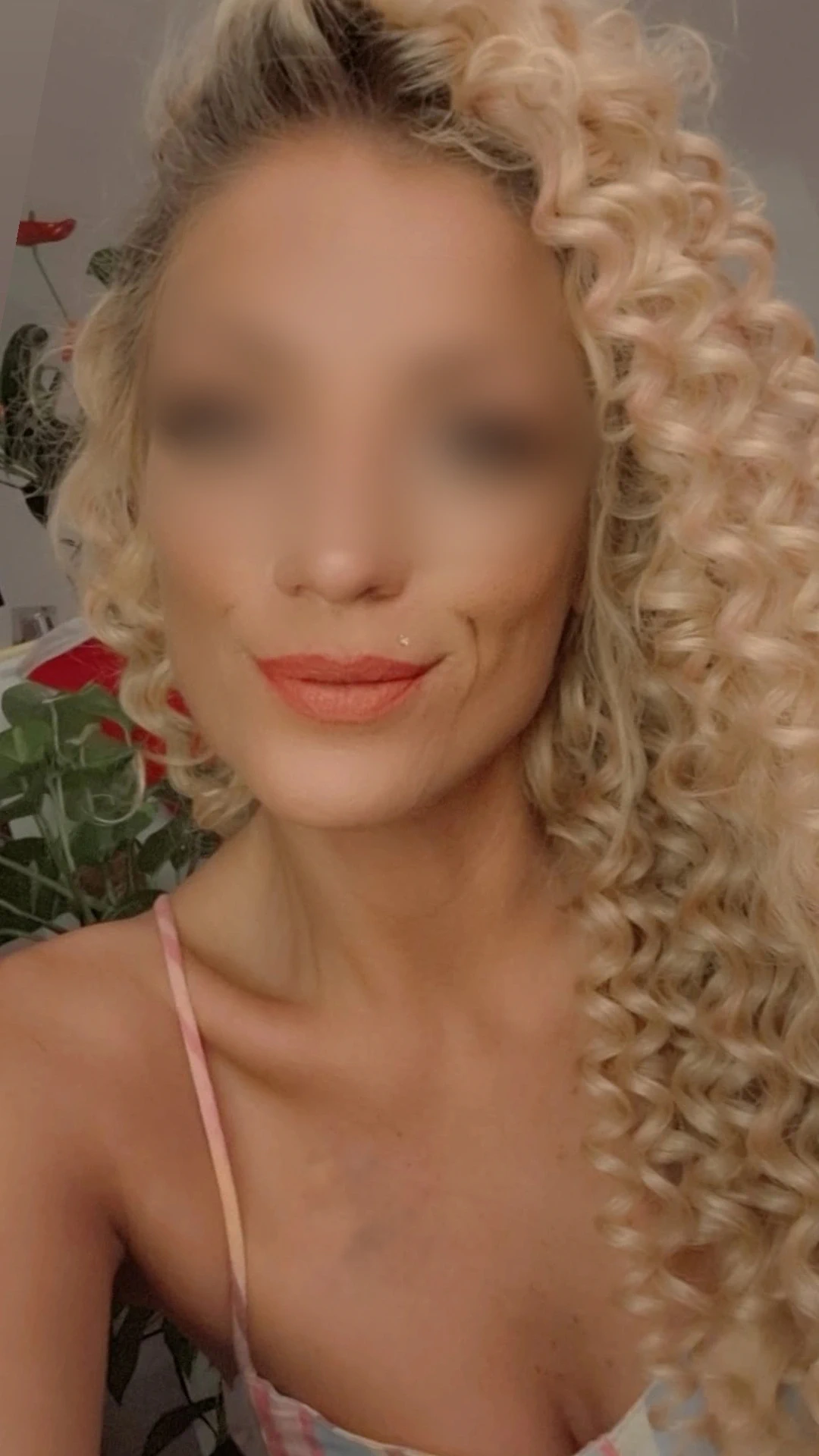 Eine Person mit langem, dicht gelocktem blondem Haar und heller Haut lächelt leicht und trägt ein hellrosa Oberteil. Im Hintergrund sind grüne Blätter und ein Hauch von rotem Dekor zu sehen. Die Gesichtszüge sind unscharf.