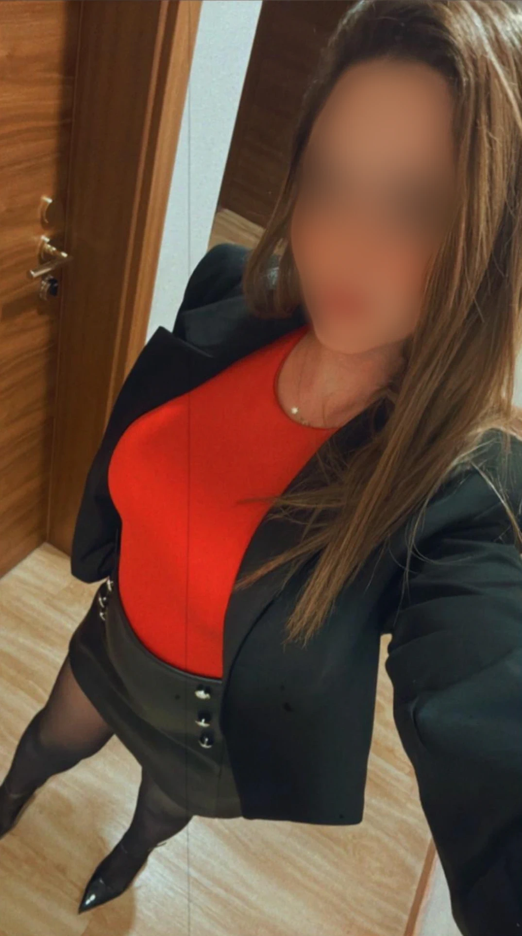 Eine Person steht in einem Haus und trägt ein rotes Oberteil, einen schwarzen Blazer, einen kurzen schwarzen Rock mit Knöpfen, eine schwarze Strumpfhose und schwarze Absätze. Ihr langes Haar liegt offen und ihr Gesicht ist unscharf. Der Schauplatz scheint ein Flur mit Holztüren zu sein.