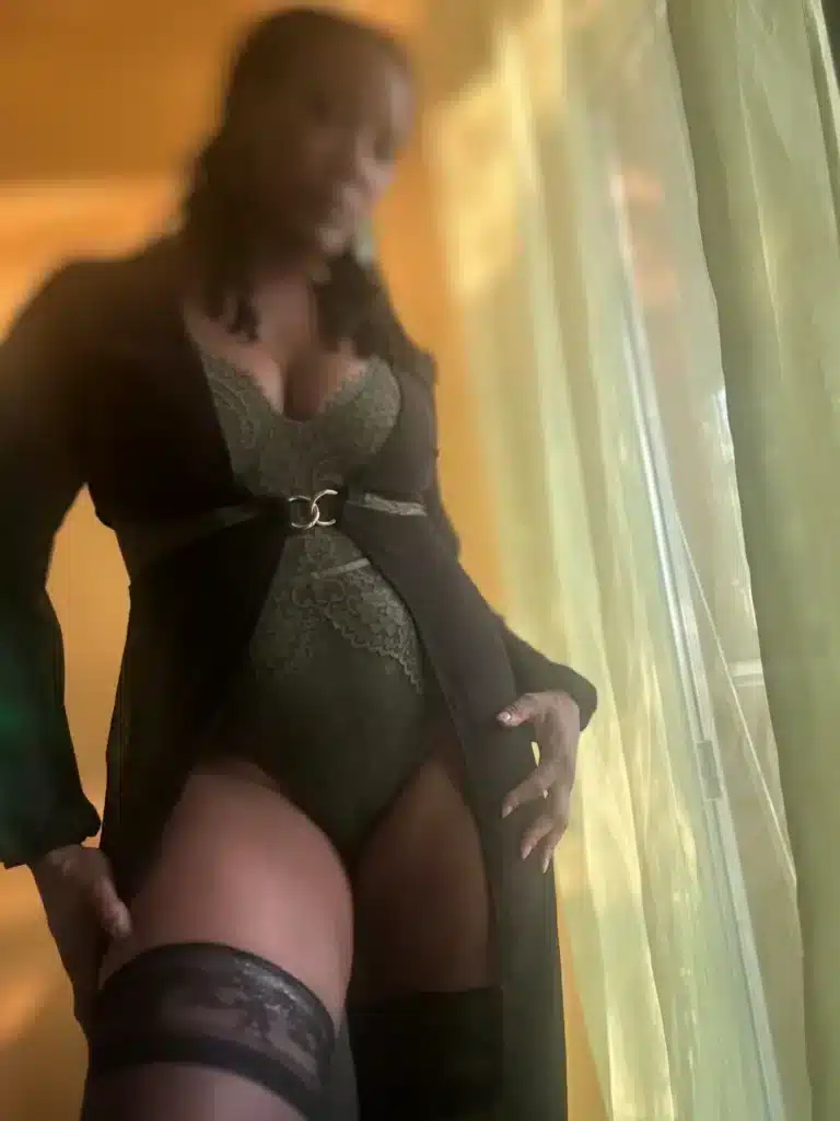 Lolaa is standing near a window with sheer green curtains, wearing lace underwear.