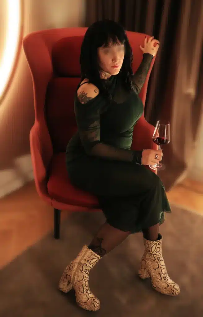 Eine Person mit dunklen Haaren und Tätowierungen sitzt auf einem modernen roten Stuhl, trägt ein dunkelgrünes Kleid und Schlangenlederstiefel und hält ein Glas Rotwein in der Hand.
