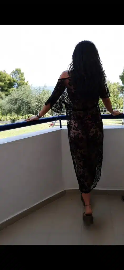 Eine Person mit langen, welligen Haaren steht auf einem Balkon, den Rücken zur Kamera gewandt, und trägt ein schwarzes Spitzenkleid. Im Hintergrund sind grüne Bäume, Gras und ein blauer Himmel in der Ferne zu sehen.