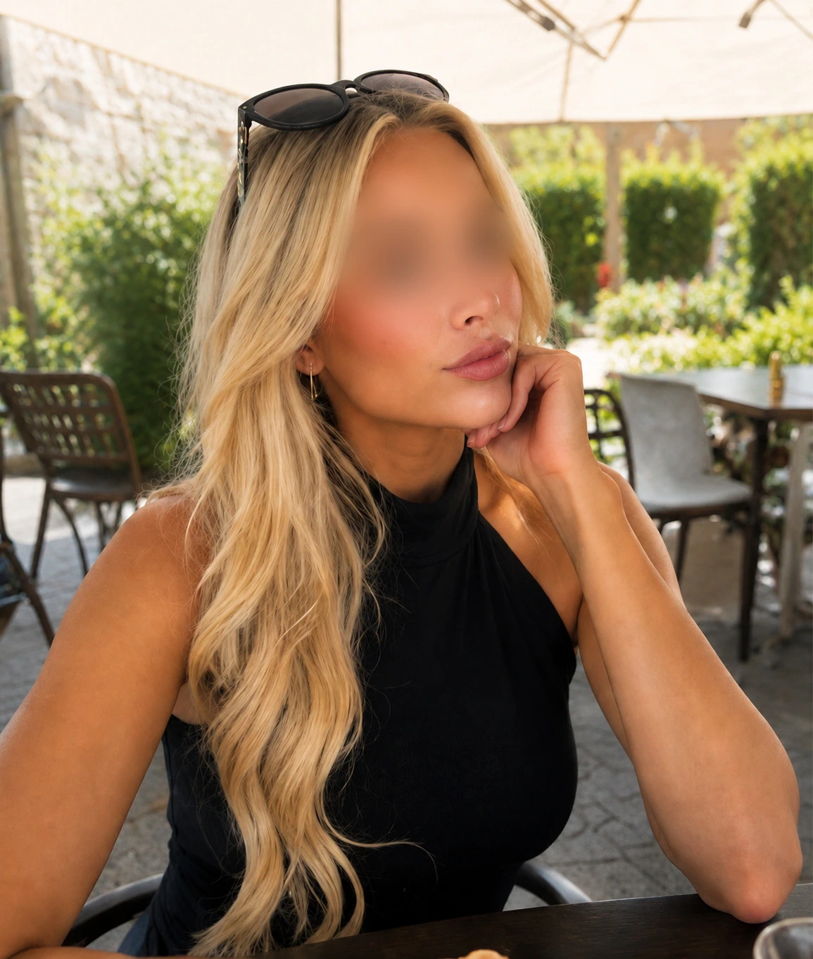 Eine Frau mit langen blonden Haaren sitzt in einem Straßencafé und stützt ihr Kinn auf ihre Hand. Sie trägt ein ärmelloses schwarzes Oberteil und eine Sonnenbrille auf dem Kopf. Im Hintergrund sind Grünflächen und Tische unter einem Sonnenschirm zu sehen.