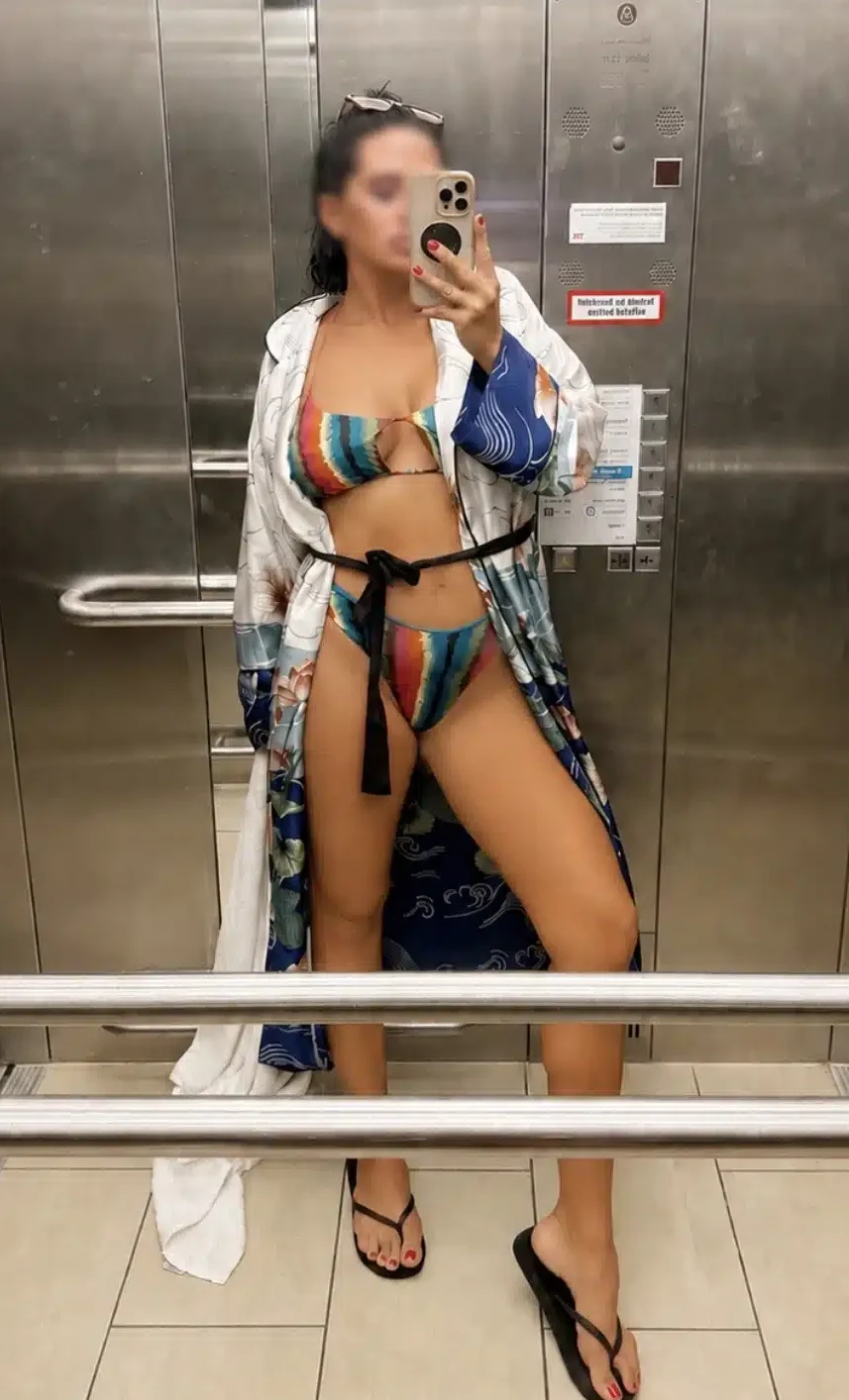 Eine Person in einem bunten Bikini, einem Bademantel und Flip-Flops macht in einem Aufzug aus Edelstahl ein Spiegel-Selfie. Aus Datenschutzgründen ist ihr Gesicht unkenntlich gemacht.
