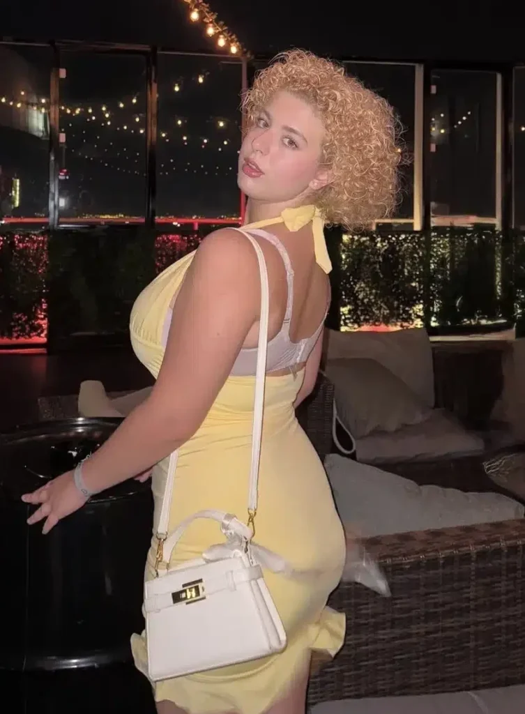 A woman with curly blonde hair, wearing a yellow dress, poses on a rooftop terrace at night; string lights hang above her, and the city lights are reflected in the glass behind her. She is holding a small white handbag and looking directly at the camera.