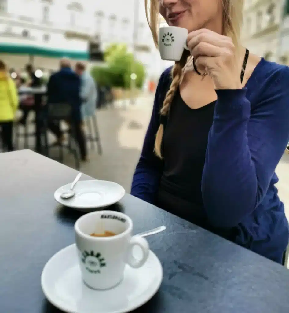 A woman with a blonde braid, wearing a dark top and a blue cardigan, is sitting at an outdoor café drinking an espresso. Another cup and saucer are on the table. The background is slightly out of focus.
