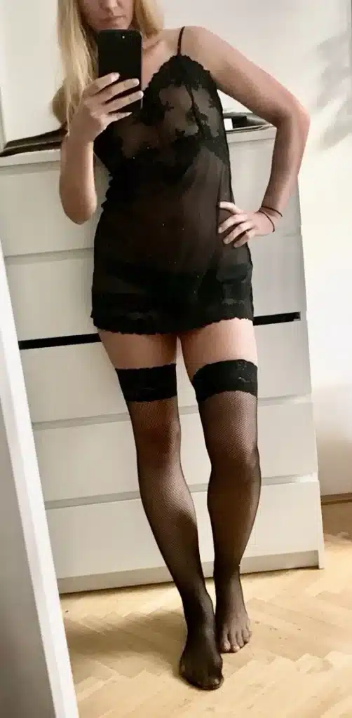 A person wearing a sheer black lingerie dress and hold-up stockings is taking a mirror selfie while standing in a room with light-colored wooden floors; a white dresser can be seen in the background.