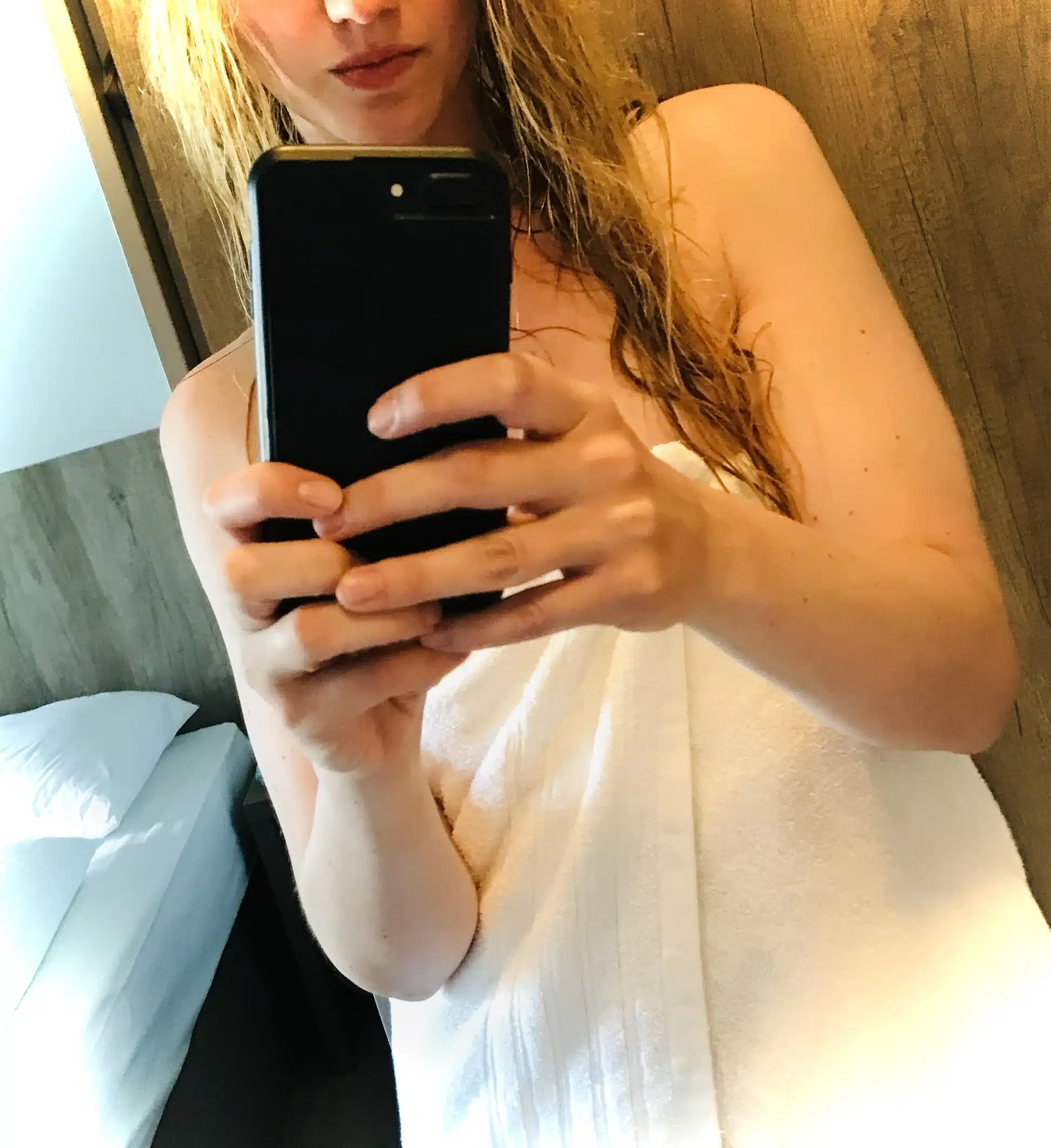 Eine Person mit nassen Haaren, in ein weißes Handtuch gewickelt, macht ein Spiegel-Selfie in einem Raum mit einer Holzwand, im Hintergrund ist ein Bett zu sehen.