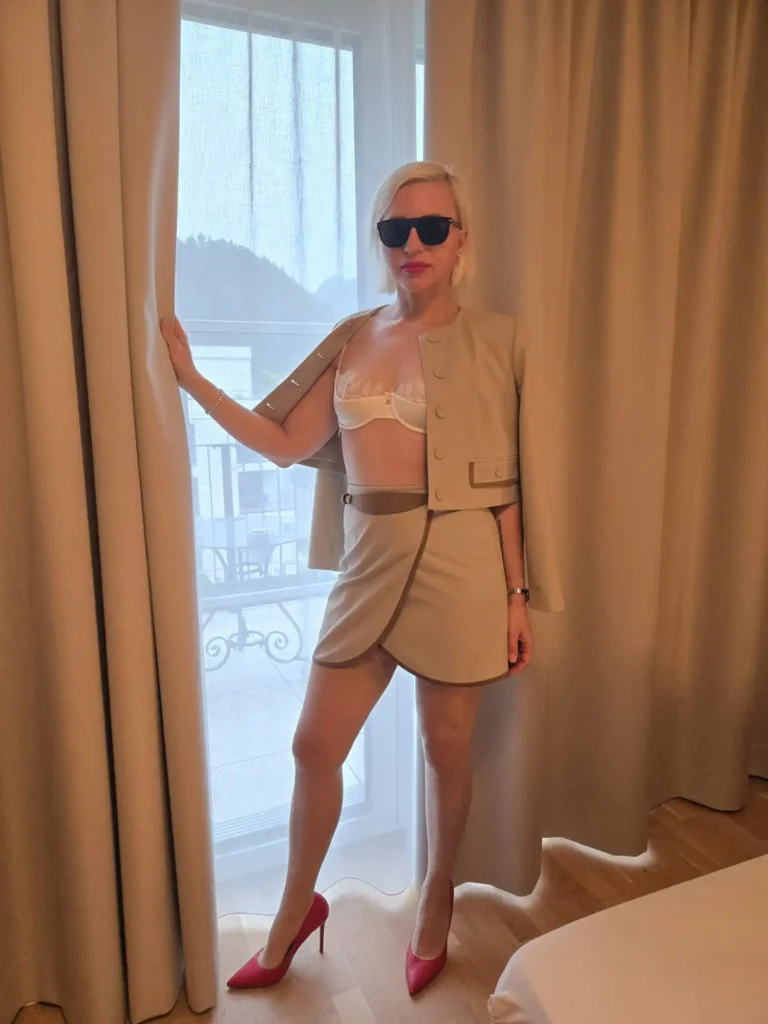 Eine Frau namens Eva, die eine Sonnenbrille, eine beige Jacke, einen Minirock, ein weißes Bralette und rosa Pumps trägt, steht an einem Fenster mit transparenten Vorhängen, wobei sie mit einer Hand den Vorhang berührt, in einem hell beleuchteten Raum mit hellem Holzboden.