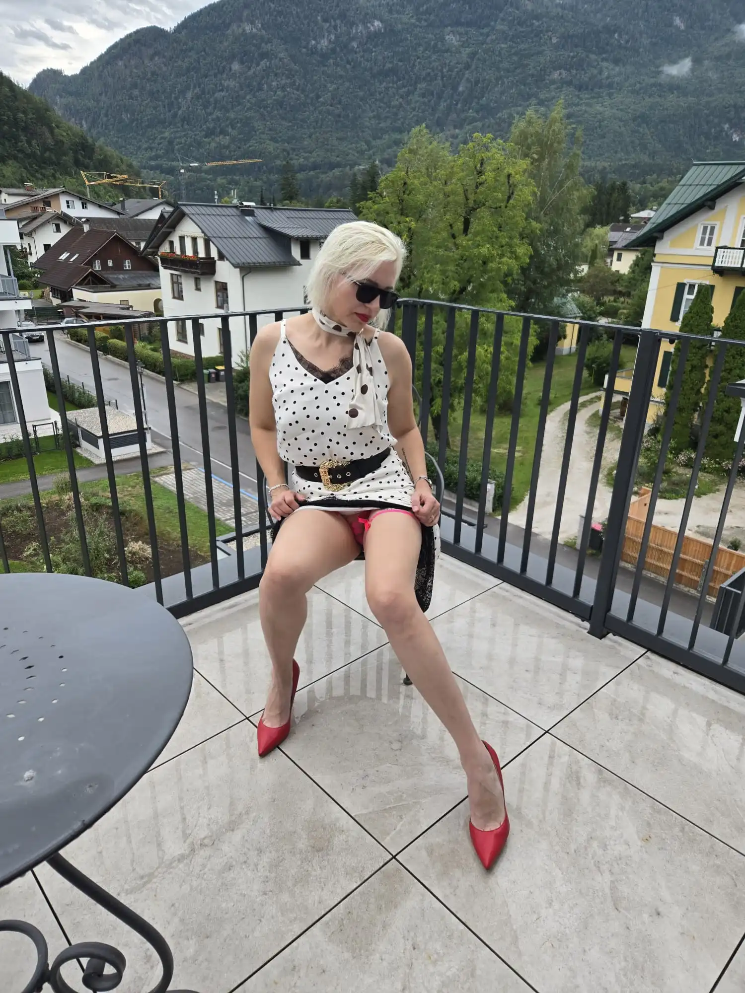 A woman named Eva, wearing a white polka-dot dress and red pumps, is sitting on a balcony chair and looking down. In the background, you can see houses, trees, and mountains; in the foreground, there is a round table.