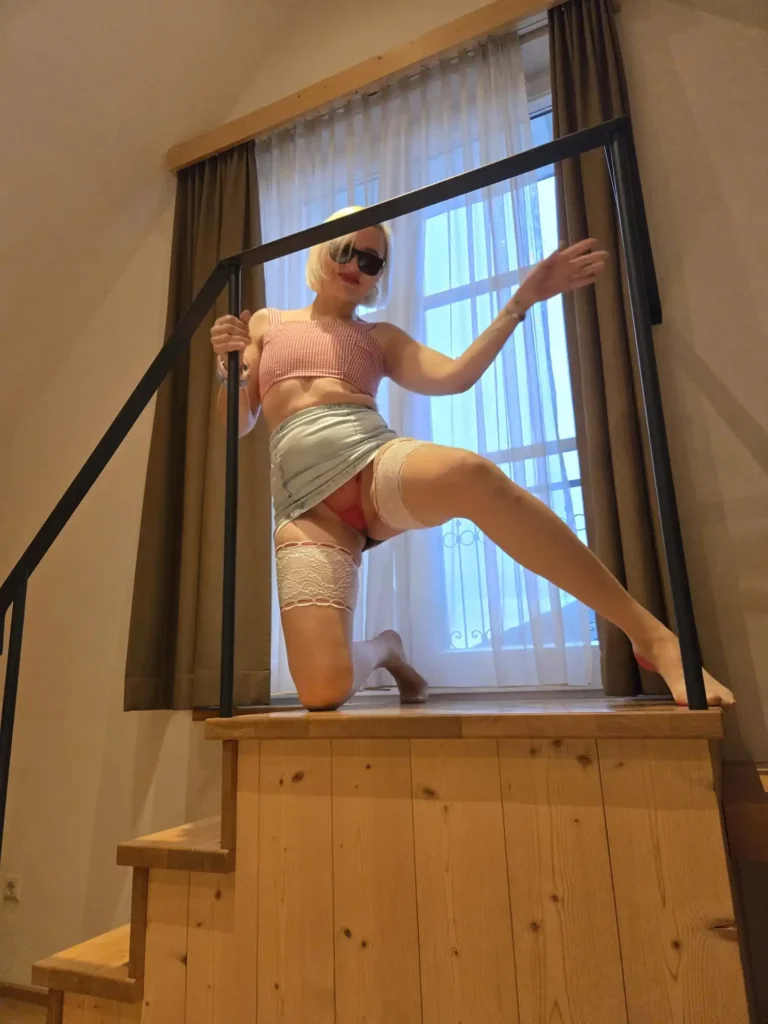 A person wearing sunglasses and blonde hair, dressed in lingerie, poses confidently on a wooden staircase in front of a window with sheer curtains, holding onto a black railing.