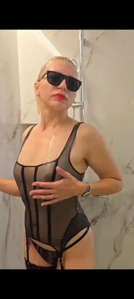Eva, die eine Sonnenbrille und einen schwarzen, durchsichtigen Body trägt, steht in einer mit Marmorfliesen ausgekleideten Dusche, aus der Wasser fließt, und posiert mit einer Hand auf der Brust.