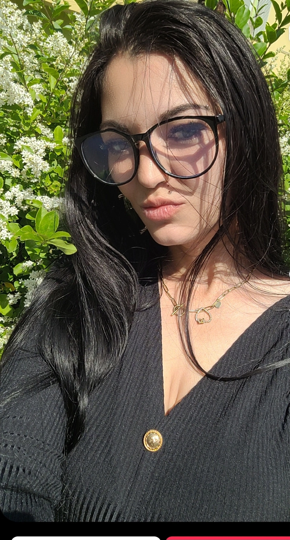 Eine Frau mit langen schwarzen Haaren und Brille macht ein Selfie im Freien. Sie trägt ein schwarzes Oberteil mit einem goldenen Knopf und eine rubinrote Halskette und steht an einem sonnigen Tag vor grünem Laub und weißen Blumen.
