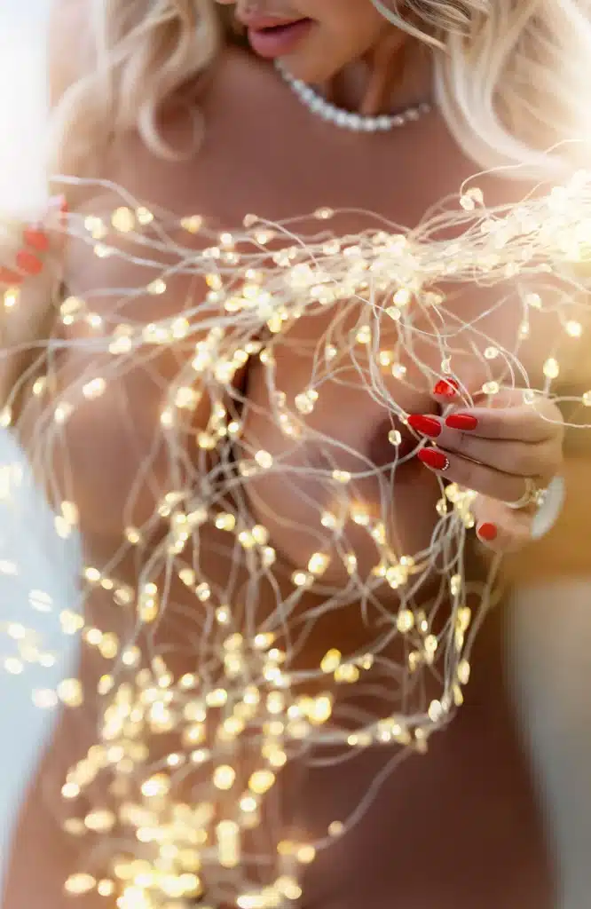 Eine Frau mit langen blonden Haaren hält leuchtende Lichterketten in den Händen und bedeckt dabei mit der Hand ihre Brust. Sie hat rote Fingernägel und trägt eine Perlenkette. Das Bild ist leicht unscharf und warm beleuchtet.